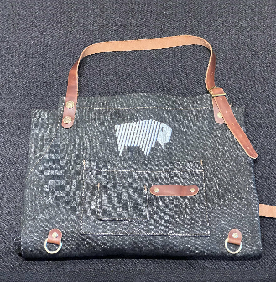 Folded denim apron with pockets and leather straps, ideal for BBQ and cooking.