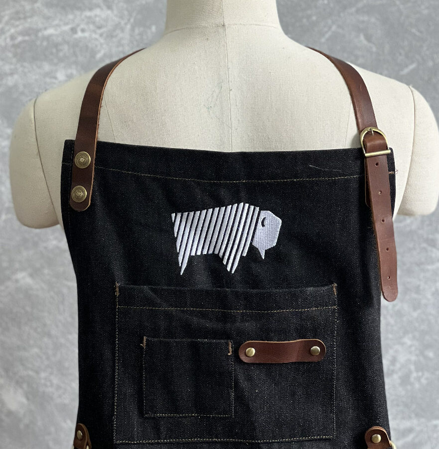 Denim apron displayed on a mannequin with an embroidered bison logo on the chest.