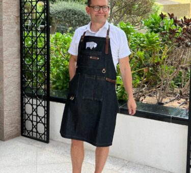 Man wearing a denim BBQ apron with multiple pockets and leather straps in an outdoor setting.