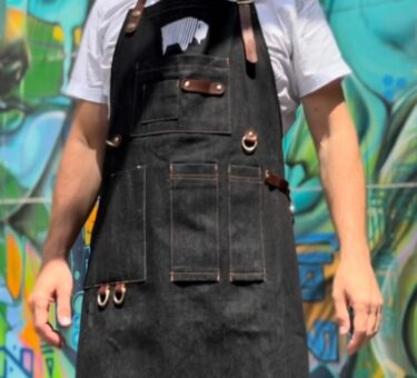 Close-up of a man wearing a denim BBQ apron with multiple pockets in front of a graffiti wall.