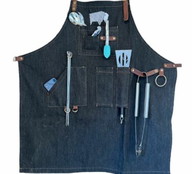 Denim BBQ apron with grilling tools displayed in multiple pockets and leather straps.
