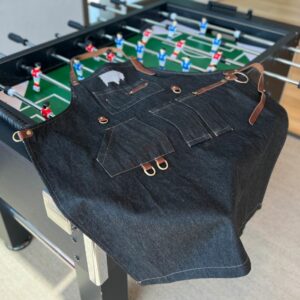 BBQ apron displayed on a foosball table in a community area, showcasing multiple pockets, a large metal ring, and a small leather belt with a brass snap fastener.
