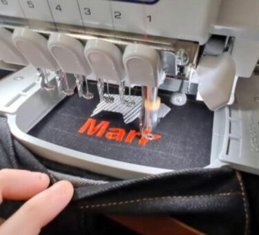 Our denim apron on an embroidery machine, being stitched with the custom text "Mark" in red thread.