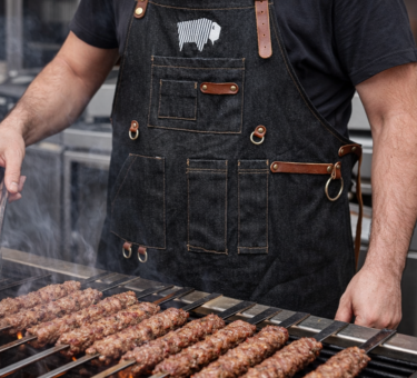 Black leather-strap BBQ apron worn while grilling Turkish-style kebabs over open flames