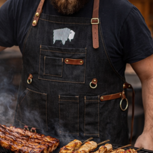 Pitmaster wearing a black BBQ apron with leather straps grilling ribs and kebabs over a smoky barbecue
