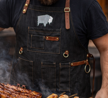 Pitmaster wearing a black BBQ apron with leather straps grilling ribs and kebabs over a smoky barbecue