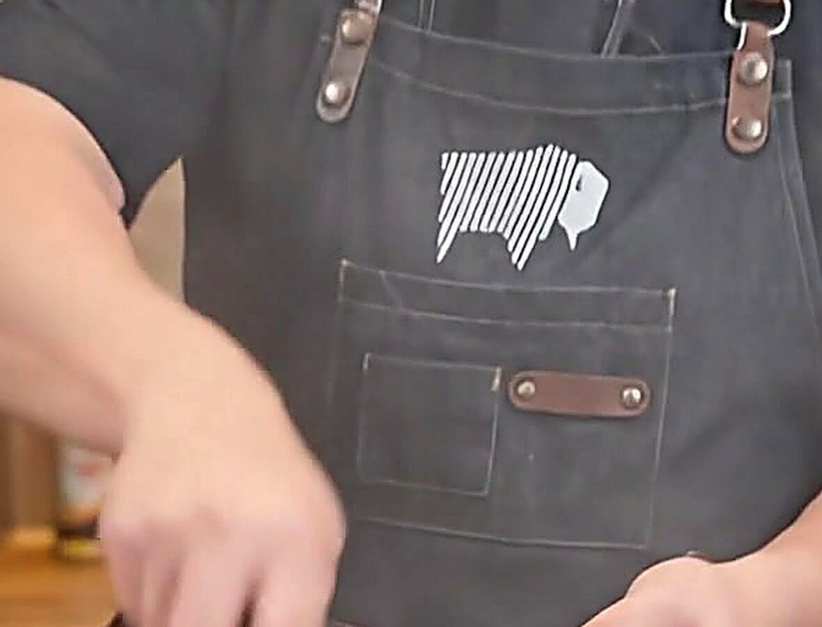 Cook wearing a black BBQ apron with leather straps using a wok over high heat in an outdoor kitchen
