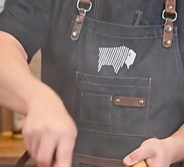 Cook wearing a black BBQ apron with leather straps using a wok over high heat in an outdoor kitchen