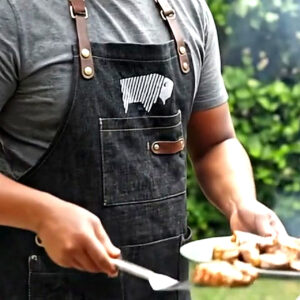 Close-up of black BBQ apron with leather trim during live-fire cooking and pan stirring