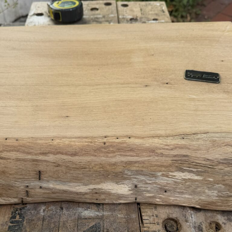 Top-down view of a thick Bison Hill Angus solid oak BBQ chopping board with a rustic live edge and branded metal plaque on a workbench.