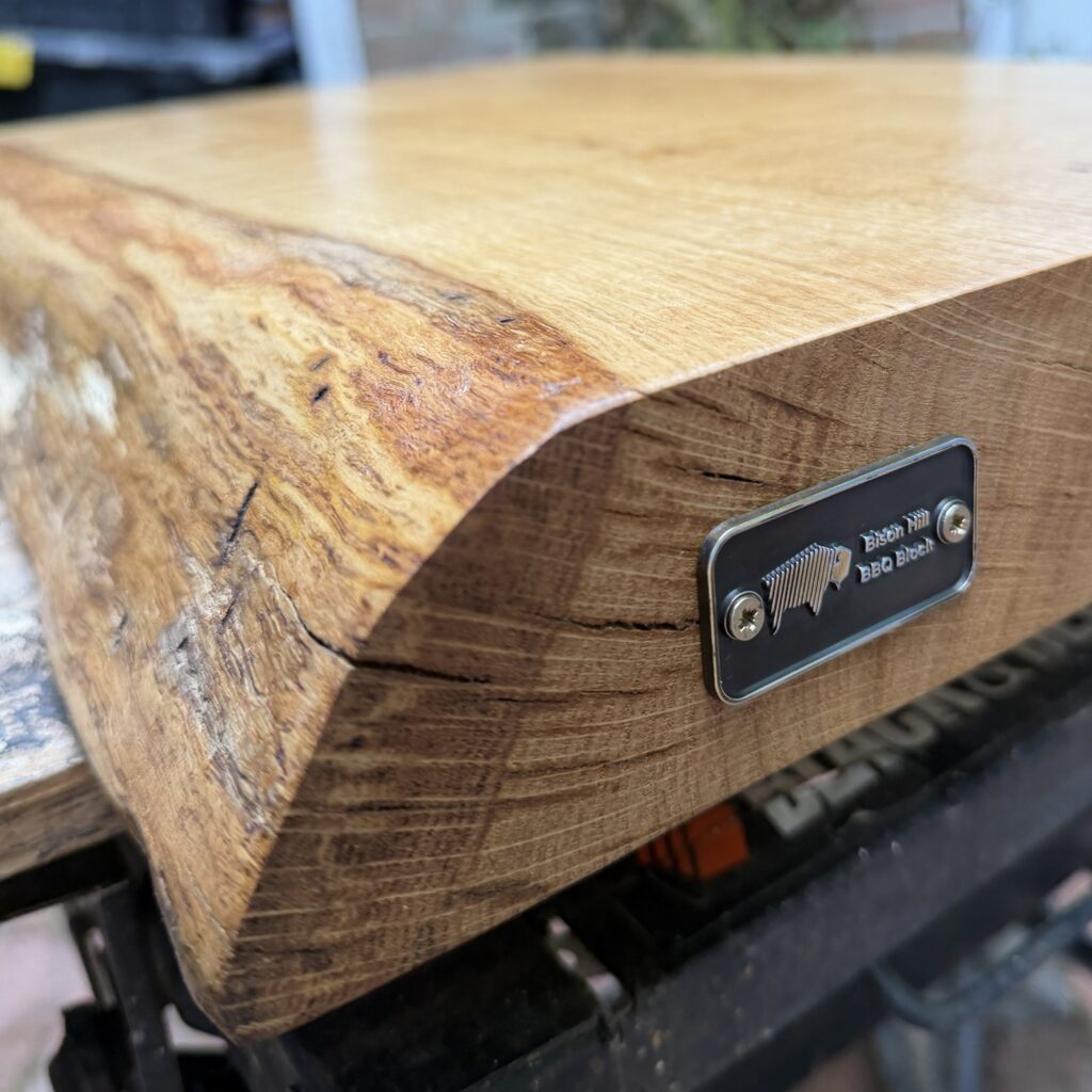 lose-up corner angle of a thick solid oak chopping board featuring a natural live-edge grain and a silver-and-black branded metal plaque screwed into the side profile.